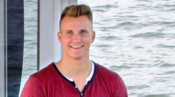 Ein junger Mann steht auf einem Boot und lächelt in die Kamera. Im Hintergrund ist Wasser.